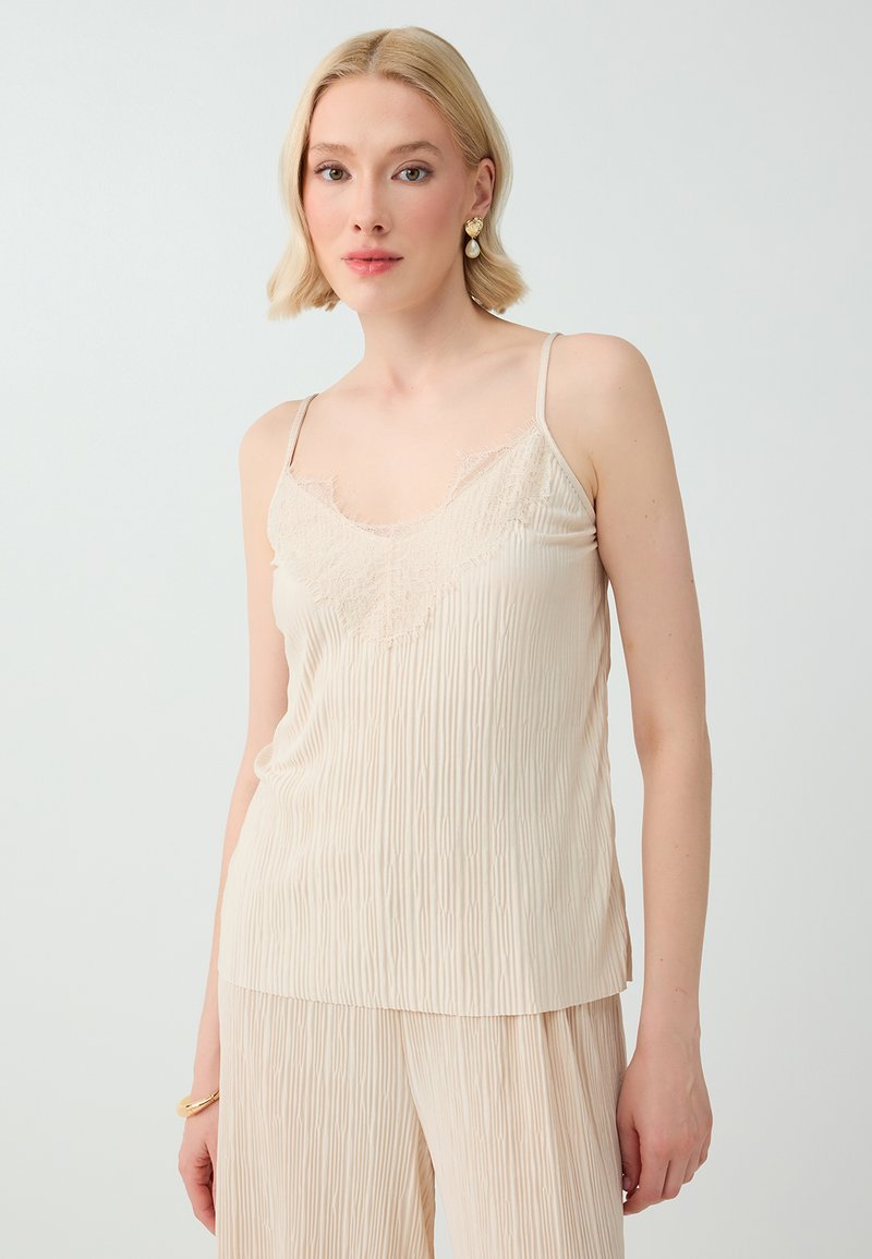 Femme aux cheveux blonds courts portant un caraco plissé crème avec une finition en dentelle et un pantalon assorti, debout devant un fond uni clair.