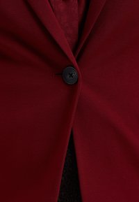 Burgundská blazerka s hladkým materiálem, jednoduchým černým knoflíkem na zapínání a jemným texturovaným detailem na vnitřním podšívce.