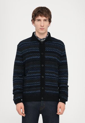 Stickad cardigan med svart bas och blåa zigzag-mönster, knappstängning och ribbade ärmslut. Tillverkad av mjukt, strukturerat tyg.