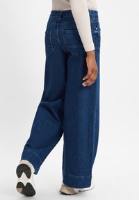 Weite, dunkelblaue Jeans mit hoher Taille, ausgestattet mit zwei Gesäßtaschen und einem sauberen Saum. Kombiniert mit beigen Turnschuhen.