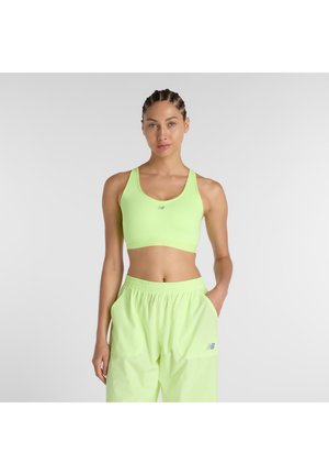 Jeune femme aux cheveux tressés, portant un soutien-gorge de sport vert néon et un pantalon ample, debout avec une main dans la poche, sur un fond uni.