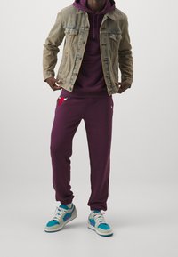 Džínová bunda přes fialovou mikinu s kapucí, v kombinaci s fialovými tepláky s červeným logem a modrými teniskami se zelenými akcenty.