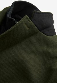 Col de manteau vert foncé avec doublure intérieure noire et fermeture éclair visible, montrant la texture et les détails du tissu superposé.