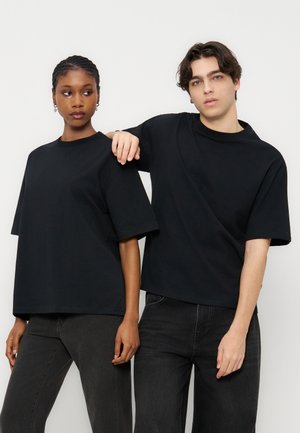 Δύο άτομα φορούν μαύρα φαρδιά μπλουζάκια και σκούρα τζιν, στέκονται δίπλα δίπλα μπροστά σε ένα απλό φωτεινό φόντο.