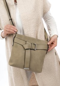 Personne portant des vêtements texturés blancs et beiges tenant un sac bandoulière taupe avec une large sangle décorative et une boucle argentée.