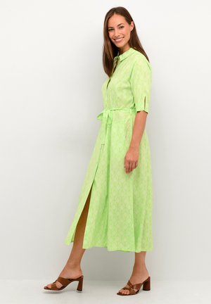 Abito camicia verde di lunghezza media con un motivo geometrico, maniche corte, cintura in vita e uno spacco laterale. Indossato con sandali con tacco marroni.