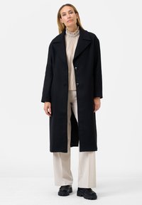 Un lungo cappotto nero con colletto a punte, design doppiopetto e tessuto testurizzato, abbinato a un top chiaro plissettato e pantaloni beige.