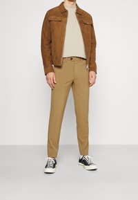 Hnědá semišová bunda se dvěma kapsami na prsou, béžové tričko a úzké béžové kalhoty. Černé nízké tenisky s bílou podrážkou, viditelné tkaničky.