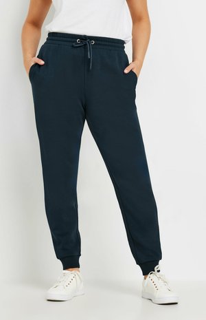 Persoon die marineblauwe joggingbroek draagt met een elastische taille en trekkoord, handen in de zakken, gecombineerd met witte sneakers en een wit topje.