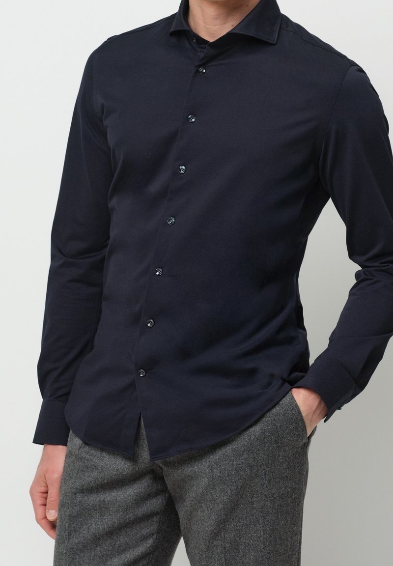 Homme portant une chemise ajustée bleu marine foncé boutonnée à manches longues et un pantalon en laine gris, une main dans la poche sur fond uni.