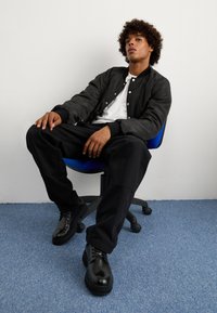 Μαύρο bomber μπουφάν με κουμπιά snap, λευκό T-shirt και μαύρο παντελόνι. Λαμπερά μαύρα παπούτσια, καθισμένος σε μια μπλε καρέ�κλα γραφείου μπροστά σε έναν λευκό τοίχο.