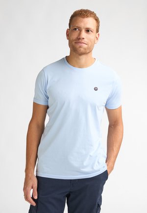Hombre con cabello rizado rojo que lleva una camiseta azul claro de cuello redondo y pantalones azul oscuro, de pie frente a un fondo blanco liso.