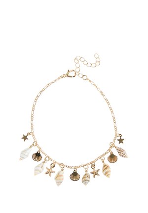 Ankle with a gold charm bracelet featuring shells and starfish, wearing brown pants and brown suede slide sandals.