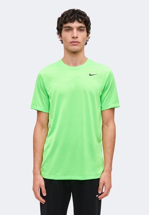 Mladi muškarac u svijetlo zelenoj Nike majici kratkih rukava i crnim hlačama stoji ispred jednobojne svijetlo sive pozadine.