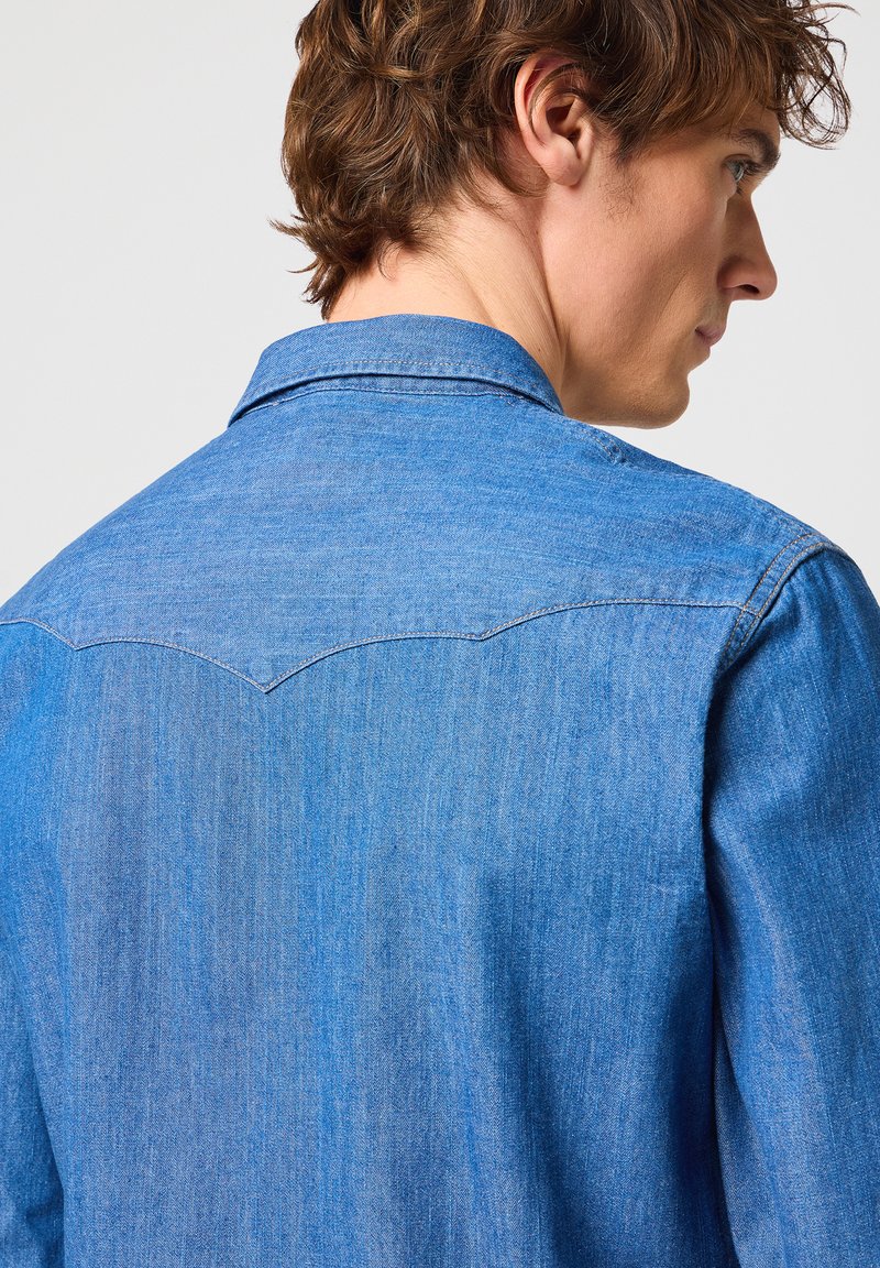 Camisa de mezclilla en azul medio, con cuello puntiagudo, detalles en el yugo y una textura suave, vista desde la parte posterior.