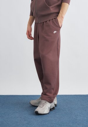 Mand iført brune Nike-sweatpants og grå Nike-sneakers står på blåt tæppe med den ene hånd i lommen.