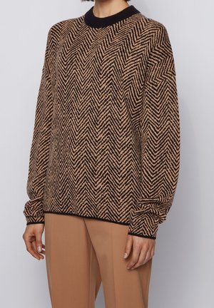 Femme portant un pull à motif zigzag marron et noir avec col et poignets noirs, associé à un pantalon couleur camel.