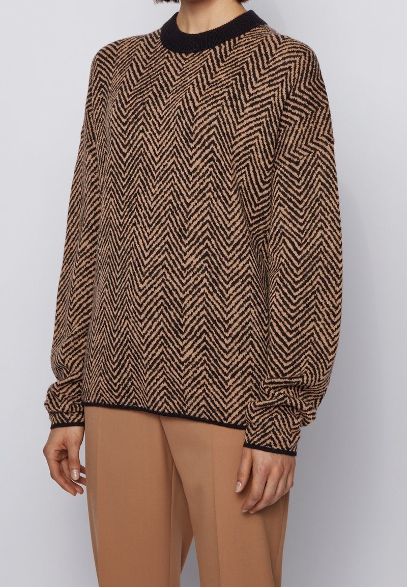 Femme portant un pull à motif zigzag marron et noir avec col et poignets noirs, associé à un pantalon couleur camel.