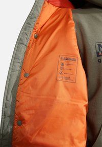 Graue gepolsterte Jacke mit orangefarbenem Innenfutter, ausgestattet mit einem Etikett, das wasserabweisenden Stoff und modulares Design beschreibt. Druckknopf im Inneren.