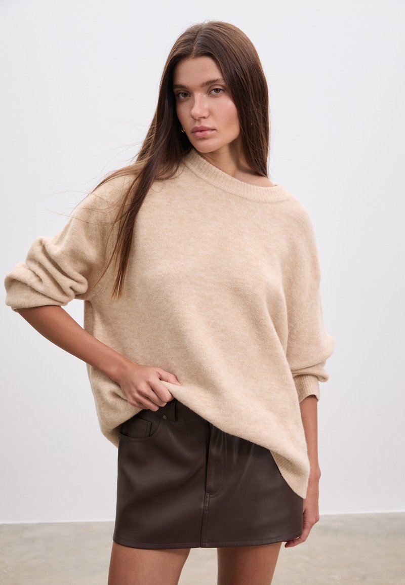 Pull beige oversize à la texture douce, col large et manches retroussées, associé à une mini-jupe en cuir marron foncé.