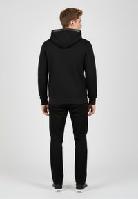 Felpa nera con cappuccio e coulisse, polsini e bordo a costine, abbinata a pantaloni neri. Il tessuto sembra morbido e resistente.