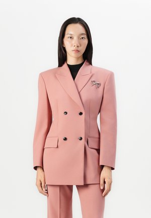 Double-breasted pink blazer in a smooth fabric, featuring black buttons and flap pockets. Includes a silver bow brooch on the lapel.