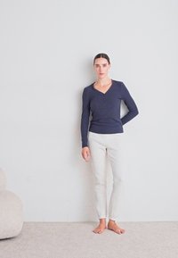 Vrouw die blootsvoets op een tapijt staat tegen een effen muur, gekleed in een marineblauw langemouwen shirt en licht gestreepte broek met haar hand achter haar rug.