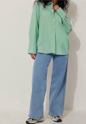 Femme portant une chemise oversize rayée verte et blanche, un jean bleu à jambes larges et des baskets blanches sur un fond clair uni.