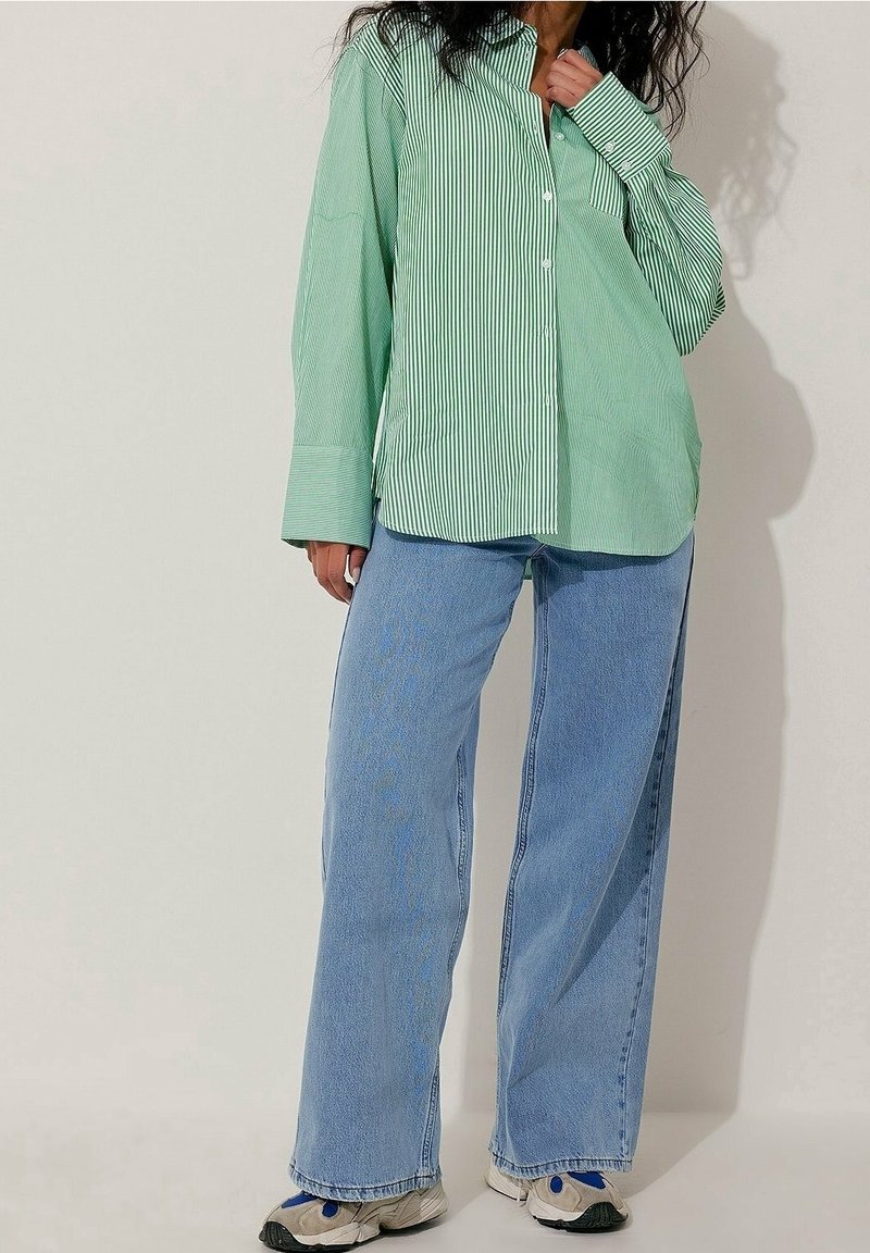 Femme portant une chemise oversize rayée verte et blanche, un jean bleu à jambes larges et des baskets blanches sur un fond clair uni.