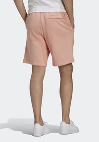 Man standing with hands in pockets wearing light pink shorts, white sneakers, and a white shirt, viewed from the back on a white background.