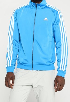 Man wearing light blue Adidas zip-up jacket with white stripes on sleeves and gray pants, standing with arms relaxed.