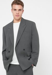 Tânăr îmbrăcat într-un sacou gri cu dungi, cu două rânduri de nasturi și pantaloni asortați, purtând un tricou alb dedesubt, cu mâinile în buzunare.