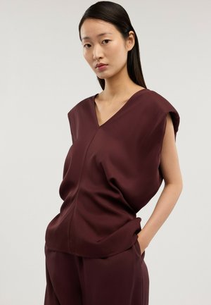 Frau mit langen schwarzen Haaren, die ein ärmelloses, dunkelbordeauxrotes V-Ausschnitt-Top und passende Hosen trägt, steht mit einer Hand in der Tasche.