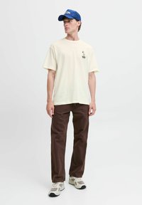 Cremefarvet bomulds T-shirt med grafisk tryk, brune lige ben bukser og grå sportsko, toppet med en blå kasket. Afslappet outfit.