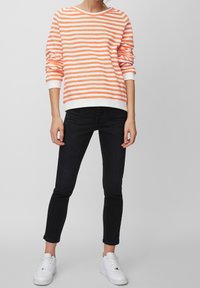 Oranssi-valkoraidallinen pitkähihainen neulepusero, jossa pyöreä pääntie ja resorit hihansuissa, yhdistetty mustiin kapealahkeisiin farkkuihin ja valkoisiin lenkkareihin.