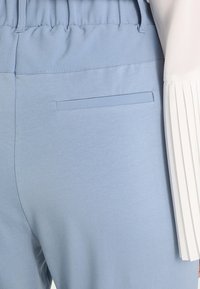 Un pantalon bleu clair avec une taille élastique, présentant une poche arrière unique et une texture lisse. Accentué par un haut blanc plissé.