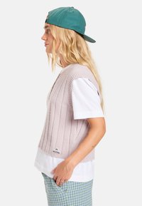 Pull à col rond sans manches, rose pâle et côtelé, porté sur un t-shirt blanc, associé à un pantalon à carreaux bleus ; portant une casquette bleu-vert. Détail du logo en argent au bas.
