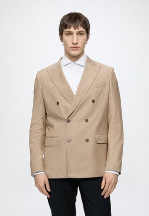 Jeune homme portant un blazer beige croisé sur une chemise blanche et un pantalon noir, debout devant un fond blanc uni.