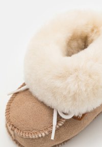 Tan semskinnslipper med en fluffy kremfarget pelskant, hvite lisser og beige søm langs sålen, som fremhever mykhet og komfort.