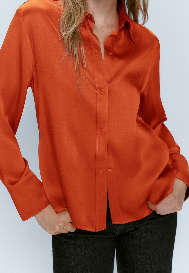 Femme portant un chemisier orange à manches longues boutonné avec de larges poignets, associé à un pantalon foncé sur un fond clair.