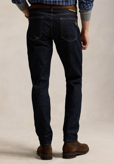 Férfi sötétkék farmerben, barna övvel, barna velúr csizmában és szürke mandzsettás kockás ingben áll, karjai nyugodtan oldalán.