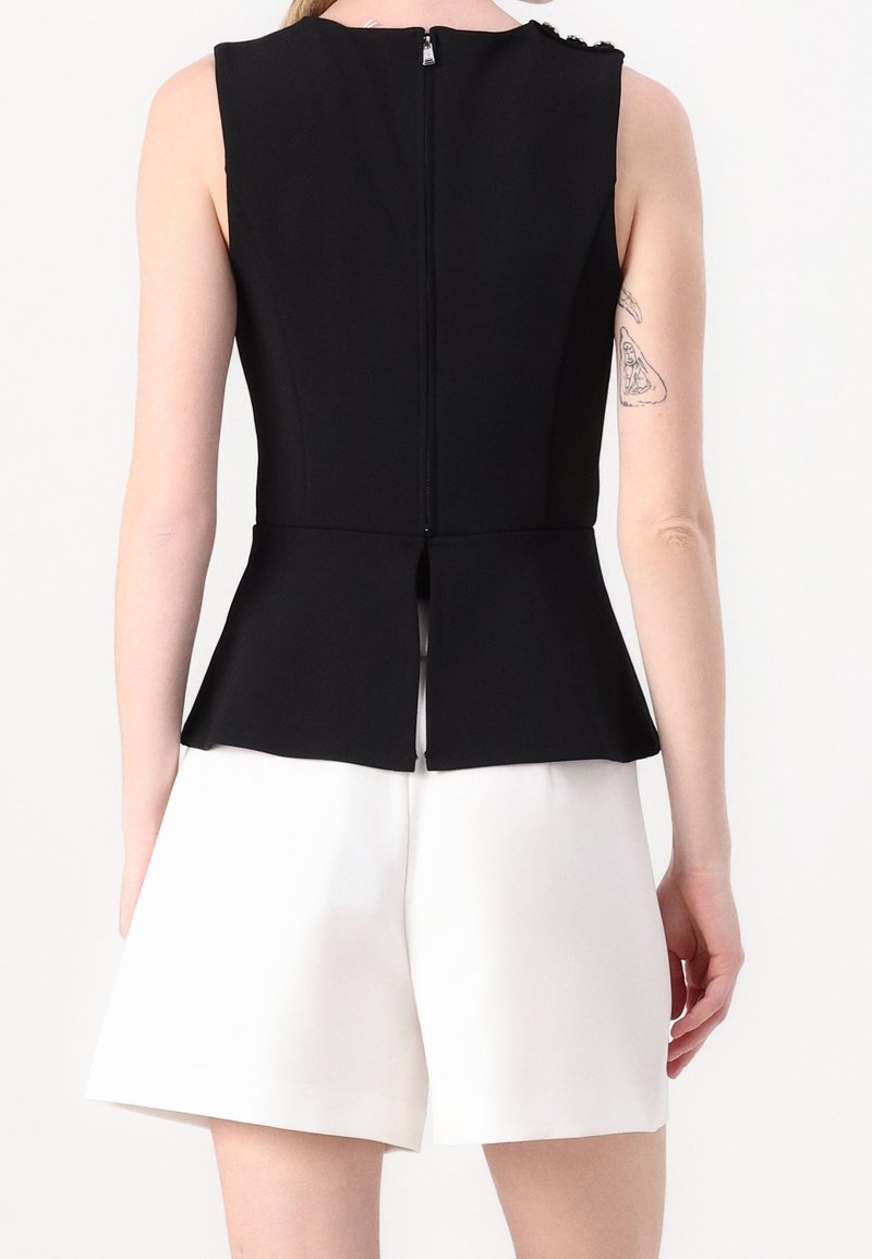Haut noir sans manches avec fermeture éclair au dos et ourlet fendu, porté avec un short blanc taille haute sur une personne debout avec un tatouage au bras.