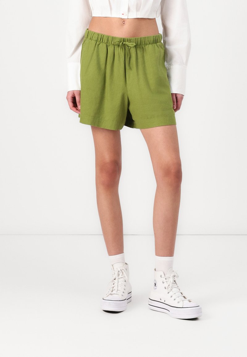 Shorts verdes con cintura elástica en un corte relajado, hechos de un tejido ligero. Combinados con zapatillas blancas de caña alta y calcetines hasta el tobillo.
