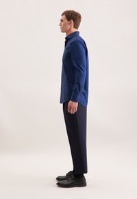 Marineblauwe overhemd met lange mouwen en een button-down kraag, gecombineerd met donkere broeken en zwarte schoenen, gepresenteerd in een zijaanzicht.