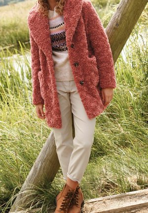 Personne portant un manteau rose chaud et texturé, un pull à motifs, un pantalon beige et des bottes marron à lacets, debout à l'extérieur près de poteaux en bois et d'herbe.