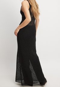 Donna che indossa un lungo vestito in pizzo nero senza maniche con un motivo a diamante, in piedi di lato su uno sfondo semplice.