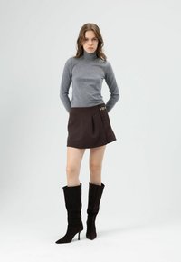 Grijze coltrui, bruine minirok met plooien en kettingdetail, gecombineerd met hoge zwarte suède stiletto laarzen. Effen achtergrond.