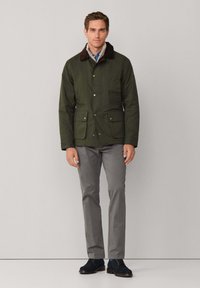 Chaqueta verde oscuro con cuello de pana marrón, cierre de botones a presión, dos bolsillos delanteros, combinada con pantalones grises y zapatos de ante azul marino.