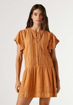 Femme portant une robe orange à manches courtes avec des boutons et des détails en dentelle, debout devant un fond clair uni.