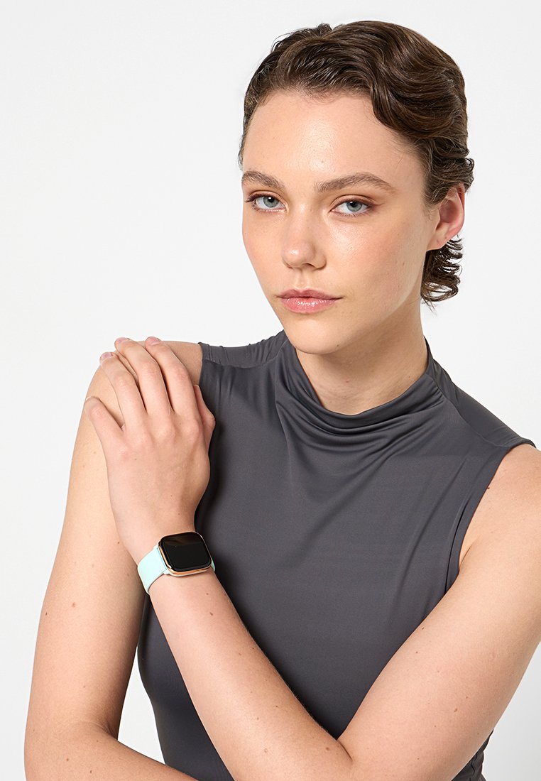 Reloj inteligente cuadrado con esfera negra, pulsera de silicona color menta, llevado en un brazo claro. El modelo lleva una blusa sin mangas gris.